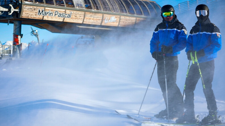 A droite, deux gendarmes sur des skis sur la neige, cette dernière vole au vent, devant des remontées mécaniques