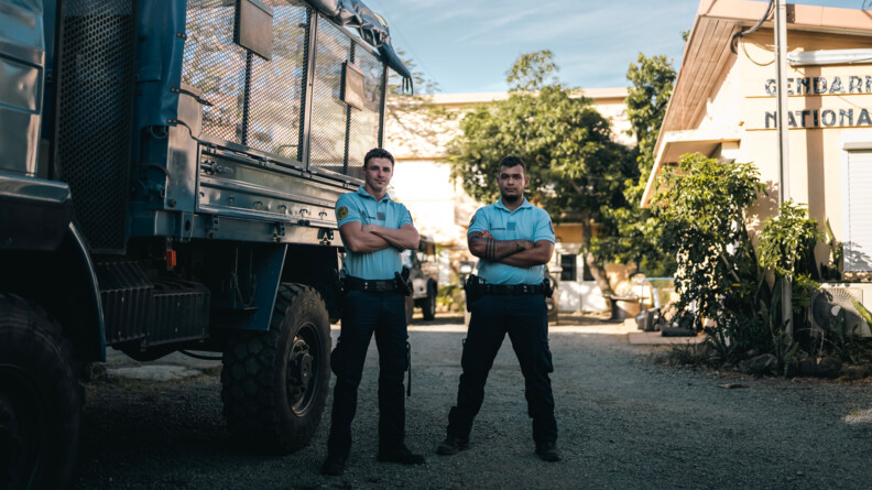 deux gendarmes debout les bras croisés, avec à droite la brigade et à gauche un camion militaire bleu