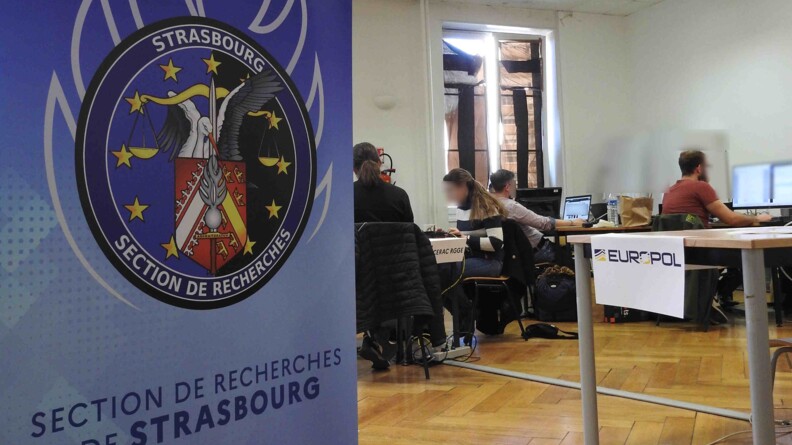 Au premier plan, un kakémono avec la rondache de la section de recherches de Strasbourg. En arrière-plan, quatre enquêteurs, de dos ou de profil, le visage flouté, travaillent derrière des ordinateurs.