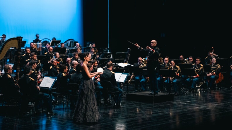 Une femme tenant un globe lumineux dans les mains avec une robe longue grise sur une scène au milieu de l'orchestre de la garde républicaine