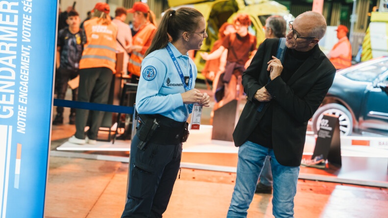 Une jeune femme gendarme, vêtue d'un pantalon bleu marine et d'un polo bleu ciel à manches longues, discute avec un visiteur sur le stand gendarmerie du Mondial de l'Auto de Paris 2024. Tous deux sont souriants.