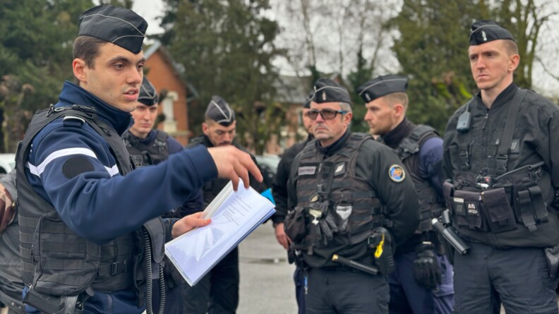 Un groupe de plusieurs gendarmes se tiennent debout dans la rue, en plein jour. L'un d'eux s'exprime, un cahier &agrave; la main. On aper&ccedil;oit en arri&egrave;re plan des arbres et la fa&ccedil;ade d'un b&acirc;timent en brique rouge.