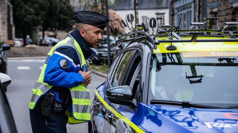 Un gendarme départemental sur le bord de la route discute avec le passager d'un véhicule d'une équipe de la course arrêté à sa hauteur.