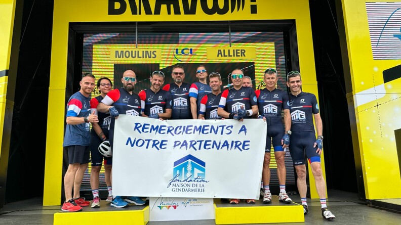 Onze cyclistes, parmi lesquels six militaires de la gendarmerie bless&eacute;s, leur encadrement et le sous-directeur de l'accompagnement du personnel, sur le podium d'arriv&eacute;e du Tour de France, avec une banderole remerciant leur partenaire, la Fondation Maison de la Gendarmerie.