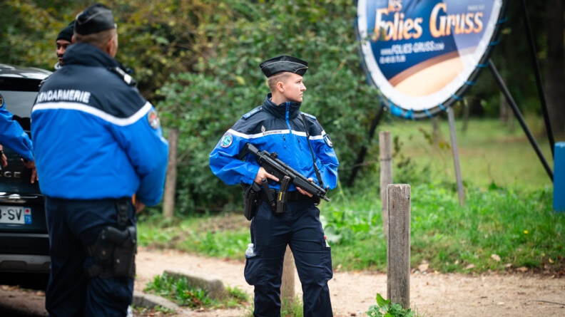 Agrandir l'image Dans un lieu boisé se tiennent debout un homme et une femme gendarmes, en tenue. L'homme est de dos. La femme, tournée vers nous, tient un fusil sur lequel sont posées ses deux mains. derrière, on aperçoit le coffre d'un véhisule sombre stationné, ains qu'une pancarte circulaire sur laquelle est inscrit en doré sur fond bleu marine la mention "Les Folies Gruss". Sont également représentés sur cette pancarte un cheval et une danseuse.