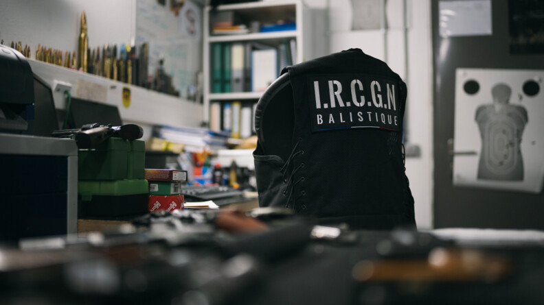 Agrandir l'image Dans une pièce illuminée par une lumière artificielle, on voit le dossier d'une chaise, recouvert d'un gilet noir sans manche portant dans le dos la mention "I.R.C.G.N Balistique, écrite en blanc, et en gros caractères. Dans le fond de la pièce, accrochée au mur, on aperçoit l'image d'une cible silhouette humaine de tir. Sur le bueau et sur le rebord du mur, on distingue un certain nombre d'objets : arme à feu, munitions de toutes tailles, boîtes, ou encore classeurs.