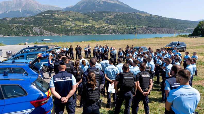 Une centaine de gendarmes sont réunis en demi-cercle autour d'un homme, également gendarme. Derrière eux, on aperçoit le lac de Serre-Ponçon, ainsi que les reliefs. Le temps ets ensoleillé. Plusieurs véhicules de gendarmerie sont stationnés autour du groupe de militaires.