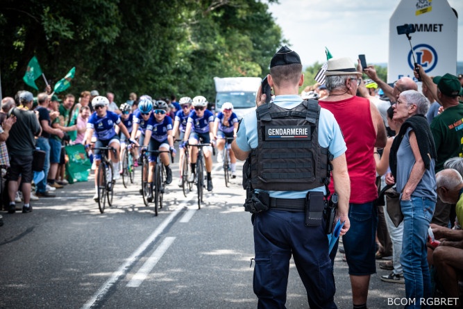 Agrandir l'image Un gendarme, vu de dos, surveille le passage d’un groupe de cyclistes féminines en tenue bleue pendant une course. Il porte un gilet pare-balles marqué « Gendarmerie ». Les spectateurs, massés des deux côtés de la route, applaudissent et prennent des photos. L’ambiance est animée et festive, dans un cadre verdoyant.