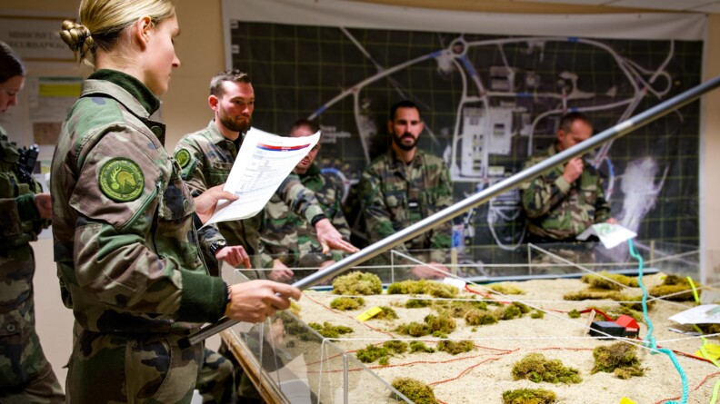 Une cavalière de la garde républicaine, de profil, en treillis, se tient debout devant la maquette d'un terrain d'opérations. Elle tient un document dans sa main gauche et une perche métallique dans la main droite. En arrière-plan, quatre autres militaires en treillis l'écoutent.