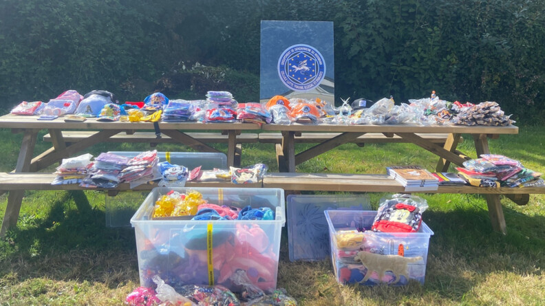 sur une table en bois avec des bancs, posée sur l'herbe, de ombreuses saisie de gendarmerie exposées dans des sacs plastiques contenant des objets, t-short,sacs etc, aux effiges de personnages deDisney, Pokemn, etc. Au sol devant le banc, deux caisses transparentes contenant le même type d'objets