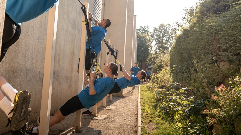 Quatre élèves gendarmes en cyclistes noirs et t-shirts bleus font des tractions devant le mur extérieur d'un bâtiment ; les deux garçons sont sur la barre et les deux filles avec des sangles.