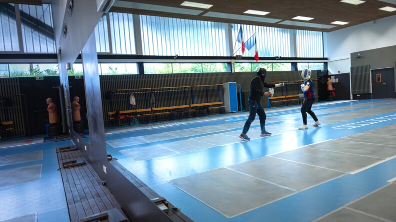 Dans le reflet d'un miroir mural deux escrimeurs s'affrontent et l'un pare l'assaut