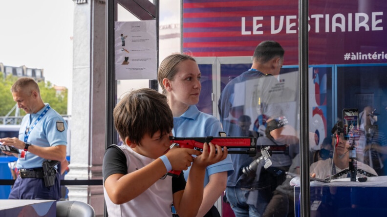 Agrandir l'image Une enfant vêtu d'un t-shirt blanc à manches noires, tire avec une carabine orange sur un stand de tir. A côté de lui se tient une femme gendarme.