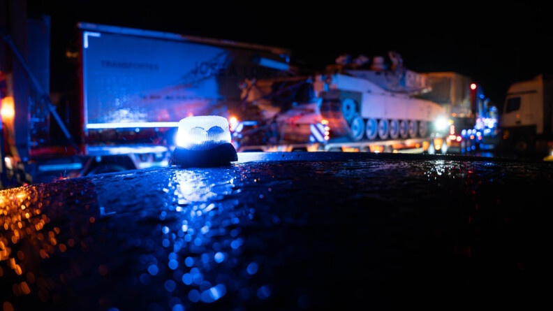 En arrière plan le convoi militaire, en premier plan un girophare de gendarmerie. Il fait nuit et sombre.