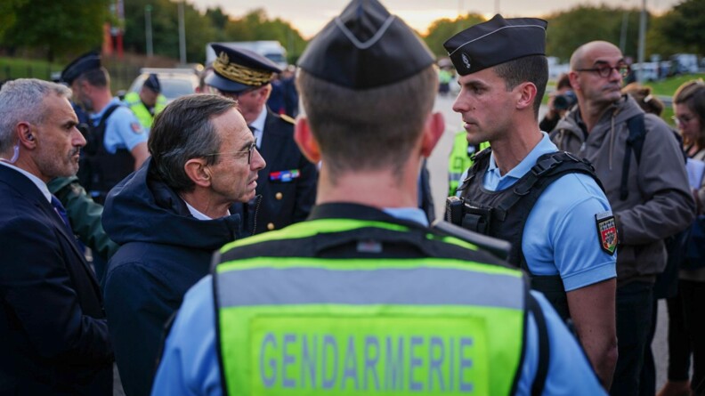 Au premier plan, un gendarme vu de dos, portant une chasuble jaune fluo avec la mention GENDARMERIE. Devant lui, &agrave; gauche, le ministre de l'Int&eacute;rieur, vu de profil, parle &agrave; un gendarme, &agrave; droite, vu de profil. Autour d'eux d'autres personnes en civil.