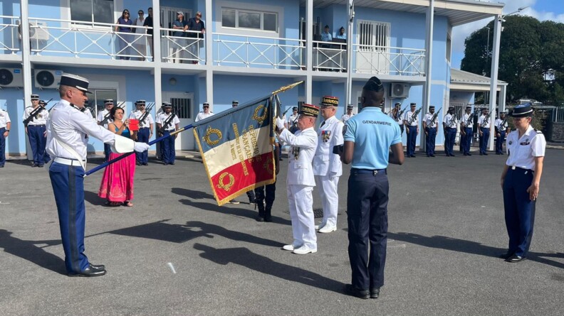Le major g&eacute;n&eacute;ral de la gendarmerie en grande tenue blanche accroche une d&eacute;coration sur un drapeau pr&eacute;sent&eacute; devant lui par un gendarme en pantalon bleu et chemise blanche. Des troupes en armes sont au garde-&agrave;-vous dans le fond.