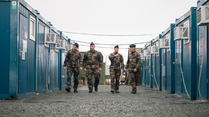 Agrandir l'image Sur une allée bordée de part et d'autres de containers bleus, deux militaires de l'armée et et deux gendarmes échangent en avançant sur les graviers
