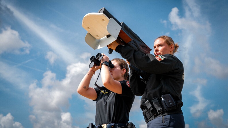 Deux femmes gendarmes, debout, côte à  côte, observent le ciel. L'une est munie d'une paire de jumelles, la seconde d'un fusil brouilleur. Le ciel est bleu et constellé de nuages blancs et gris.