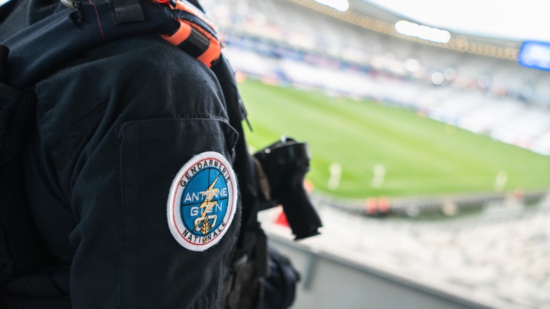 Vue sur un avant-bras vêtu de noir portant la rondache de l'Antene GIGN de la gendarmerie nationale. en bas, flou, un stade vide