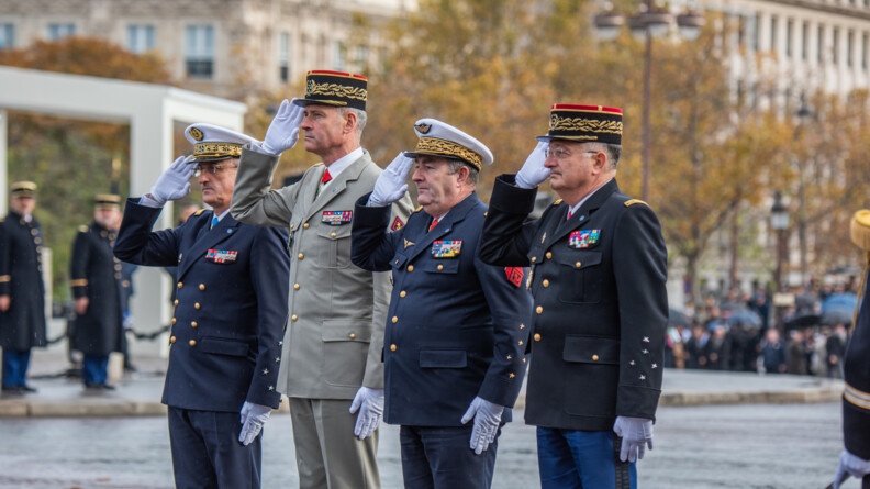 Quatre militaires, trois en tenue sombre, le quatrième vêtu d'une tenue beige, se tiennent deboit côte à côte. Leur bras gauche est positionné le long de leur cuisse gauche, tandis qu'ils effectuent un salut de la main droite. Derrière eux, on apercçoit des arbres dont les feuilles sont aux couleurs de l'automne, et la façade de bâtiments en pierre.