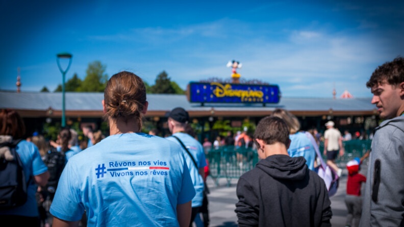 Vue de dos, en buste, des familles vêtues d'un t-shirt bleu clair #Rêvonsnosviesvivonsnosrêves, floqués d'une ligne bleu blanc rouge, se dirigeant vers le portail d'accueil de Disney
