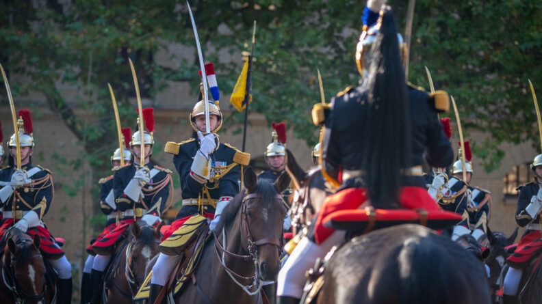Au premier plan  flou, un cavalier de la Gade républicaine de dos, à cheval. Face à lui, un peloton de la Garde présente le sabre
