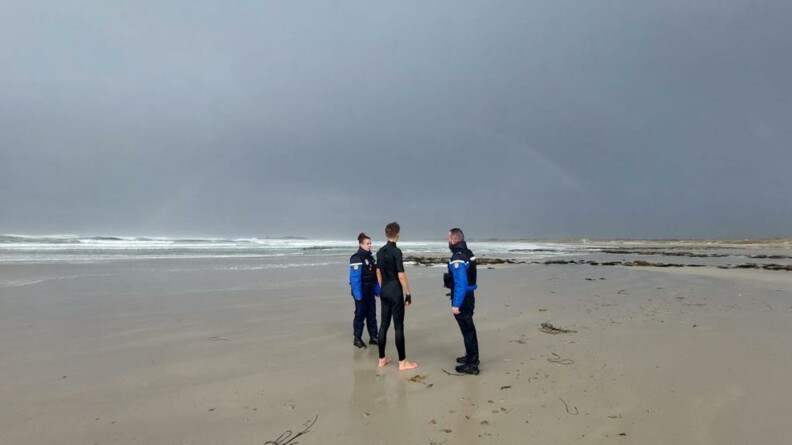 Sur une grande plage déserte, sous un ciel gris, deux gendarmes en tenue, discutent avec un homme en combinaison noire, que l'on aperçoit de dos