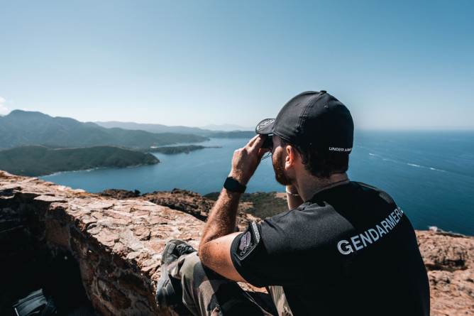 Agrandir l'image Un gendarme, assis sur un muret en pierres surplombant la mer, regarde l'horizon avec des jumelles. Le militaire se tient de dos et l'on peut lire dan sle dos de son polo noir l'inscription " Gendarmerie". il porte aussi une casquette noire et unpantalon treillis kaki. On aperçoit les montagnes bordant la mer.