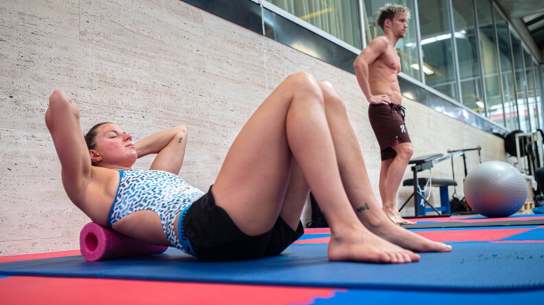 Une sportive vêtue d'un maillot de bain blanc et bleu, ainsi que d'un short noir court, réalise des exercices d'étirement, allongée sur le dos, les yeux fermés, sur un tapis de sol de couleur bleue. Sa nuque repose sur un appui tête rose.