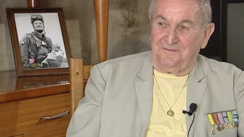 Photo du gendarme Weisse datant de 2004. Photo en buste d'un homme &acirc;g&eacute; assis sur une chaise, portant un haut jaune pale et une veste beige, sur laquelle sont pendues six m&eacute;dailles. Il porte une cha&icirc;ne en or, au bout de laquelle on voit un m&eacute;daillon. Un micro est clips&eacute; sur le revers de sa veste. Derri&egrave;re lui, sur un buffet bas en bois vernis, une photo en noir et blanc d'un soldat dans un cadre en bois.