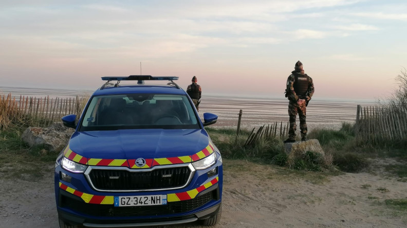 Un véhicule de gendarmerie sérigraphié, et derrière lui et et sur la droite, un gendarme, sur le litorral devant la mer