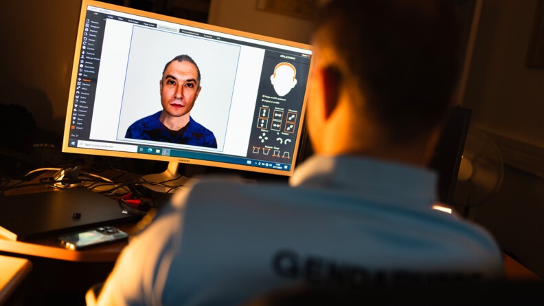 Dans une pièce sombre, on voit au pemier plan un gendarme de dos, vêtu d'un polo bleu avec l'inscription "Gendarmerie", assis devant un bureau. Face à lui, l'écran d'un ordinateur affichant le visage d'un homme d'âge moyen sur un logiciel.