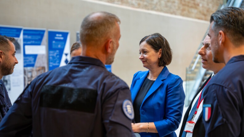 Un gendarme en combinaison bleu foncé, vu de dos, avec une rondache Frontex sur le bras droit ; à droite, deux autres gendarmes de profil, avec au premier plan l'écusson France sur le bras gauche. Au second plan, au centre, la directrice adjoint de Frontex, vêtue d'un haut noir et d'une veste bleue.