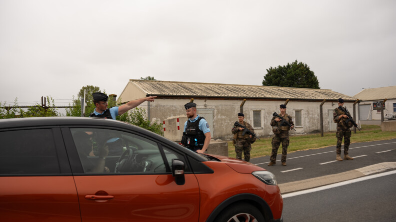 Au premier plan, un véhicule couleur rouille, et au second plan, deux gendarmes, dont l'un montre une direction au conducteur. En arrière plan, sur le bord de la route, trois militaires de Sentinelle armés de fusils. Derrière eux, une clôture haute avec des barbelés devant des baraquements.
