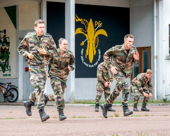 Agrandir l'image Des élèves en tenue camouflage courent côte à côte sur un sol en bitume rouge. Derrière eux, sur un mur noir, est inscrite en couleur blanche la mention : "Fort seul, mais plus fort ensemble"