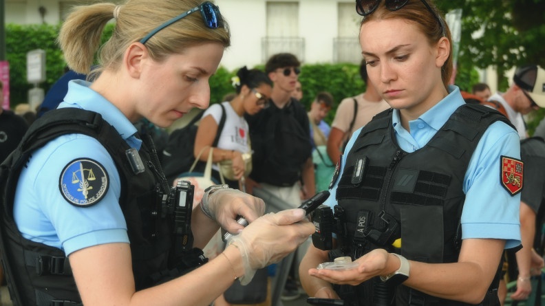 Deux jeunes femmes gendarmes se tiennent debout, l'une en face de l'autre. Les deux militaires tiennent en main leur téléphone portable professionnel. Et l'une tient une petite coupelle ronde transparente posée à plat dans sa main gauche. La scène se déroule à l'occasion du festival Garorock