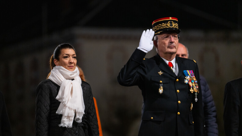 Agrandir l'image A gauche une femme en noir avec une écharpe blanche, les bras le long du corps, et le Directeur général de la gendarmerie nationale en vareuse, effectuant le salut militaire