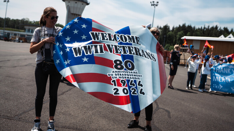 Deux femmes sur un tarmac tiennet un grand drapeau américain en forme de coeur surmonté des inscriptions en noir "WELCOME WWII VETERANS 80e anniversaire du débarquement en Normandie 1944 2024"