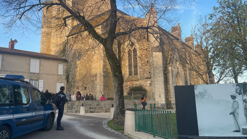 à gauche un véhicule gendarmerie, devant lui une gendarme de profil. au premier plan à droite une photo de Jean de Lattre devant un parc d'herbe. Au second lan au centre un arbre sans feuille. En arrière plan une église où se reflète le soleil