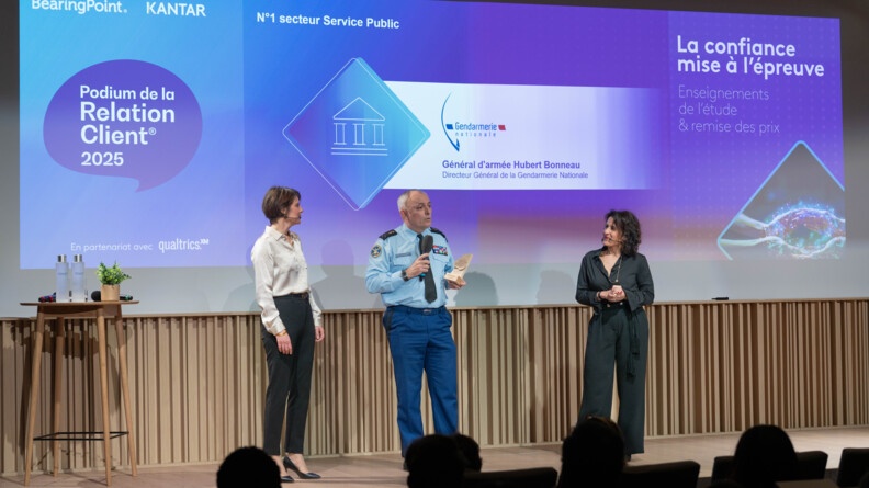 Agrandir l'image Sur une scène, un général de gendarmerie, tenant un trophée en bois et un micro, devant un écran bleu mentionnant, de gauche à droite "BearingPoint, Kantar, Podium de la relation client" , "n°1 secteur service public Gendarmerie nationale, général d'armée Hubert Bonneau, DGGN";" la confiance mise à l'épreuve"
