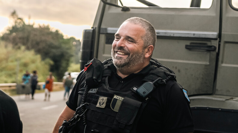 Portrait en buste d'un aum&ocirc;nier de gendarmerie regardant vers l'avant gauche, souriant. Derri&egrave;re lui, la cabine d'un camion kaki
