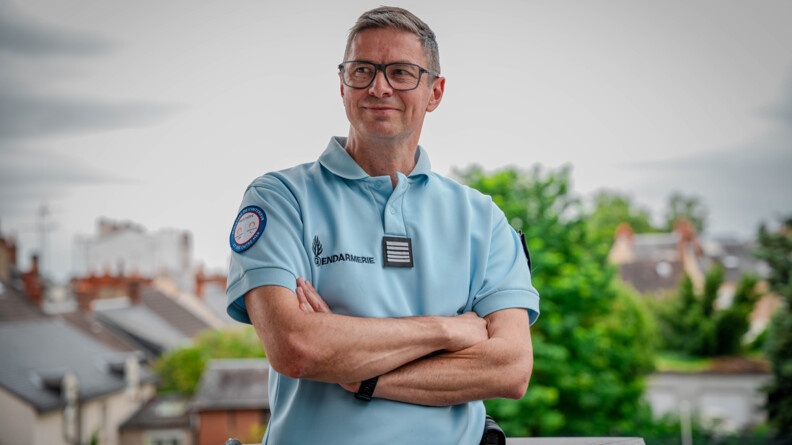 Portrait pris sur un balcon du colonel Texier en plan buste, de face, bras croisés. Il porte un polo gendarmerie bleu. En arrière-plan, des toitures et des arbres dans le flou.