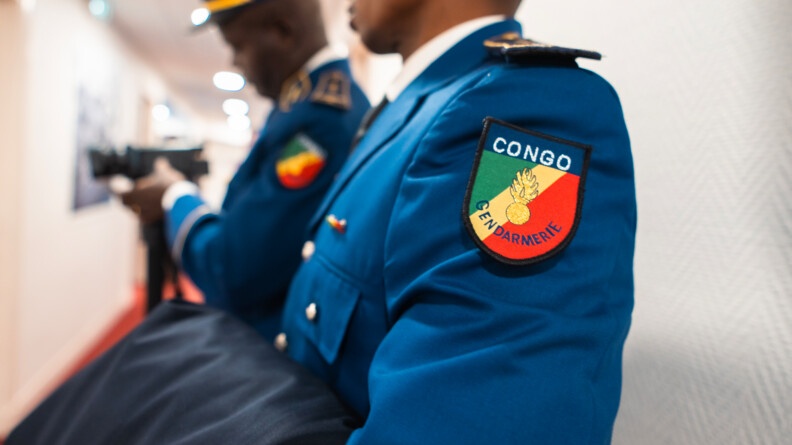 Agrandir l'image Deux gendarmes congolais se tiennent de bout de biais dans un couloir. Le second, qui est flouté, tient une caméra sur pied. Sur la manche du second, on distingue un écusson sur le haut de la manche de la veste, sur lequel est inscrit CONGO GENDARMERIE.