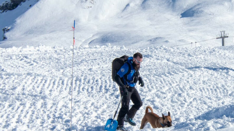 Agrandir l'image Sur une montagne enneigée, on voit une jeune homme vêtu d'une tenue de ski, équipé d'une pelle bleue, d'un gros sac à dos noir et de lunettes de ski blanches, précédé par un chien dont le museau est dirigé vers le sol.
