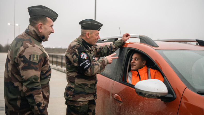 Agrandir l'image A gauche de l'image, de profil droit, deux prévôt échangent avec les militaires de l'arme du Train dans un SUV orange, et revetant un blouson orange. Le prévôt le plus près du véhicule a le bras gauche posée sur le toit du SUV et fait un signe du chiffre deux de la main droite
