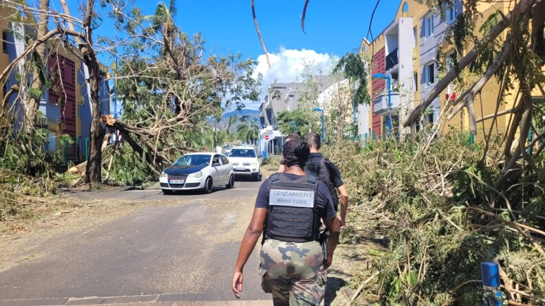 Photo de gendarmes de dos, qui évoluent sur une route, jonchée d'arbres cassés, après le passage du cyclone Garance sur l'île de la Réunion. Des voitures arrivent face à eux sur la route. Le ciel est bleu.