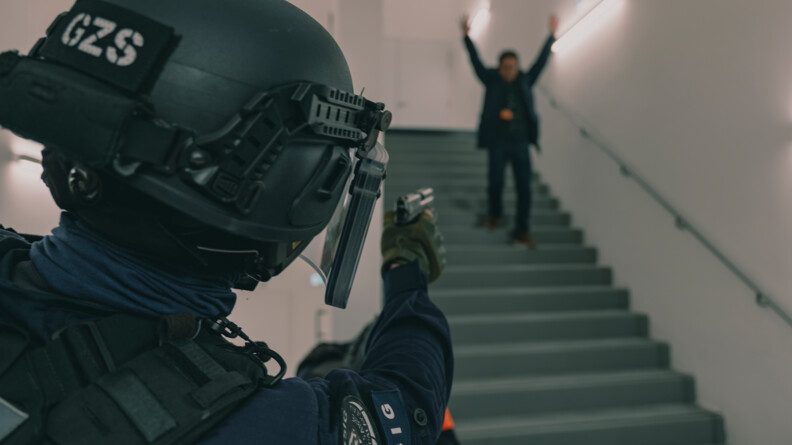 Au premier plan, un gendarme du PSIG Sabre de Palaiseau, de dos, casque sur la tête, pointe son arme en direction d'un individu qui descend les escaliers, mains en l'air.