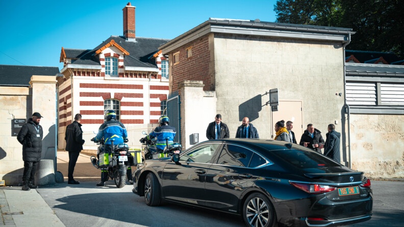 Une grosse berline noire, escortée par deux gendarmes motocyclistes, pénètrent dans l'enceinte d'un bâtiment, se présentant au niveau d'une double grille, dont les deux battants sont ouverts