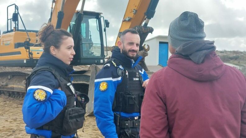 Deux gendarmes, un homme et une femme, discutent avec un homme que l'on voit de dos, vêtu d'un blouson bordeau et d'un bonnet gris. Derrière eux, se trouve un engin tractopelle, ainsi qu'une maisonnette blanche avec une porte bleue. Le ciel est gris et nuageux.