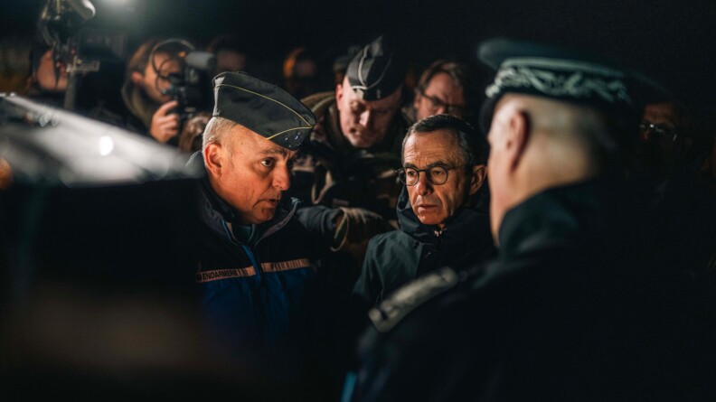 Photo du ministre de l'Intérieur, se tenant de bout aux côtés du directeur général de la gendamerie nationale. Les deux hommes sont entourés de militaires et de photographes. La scène se déroule de nuit.